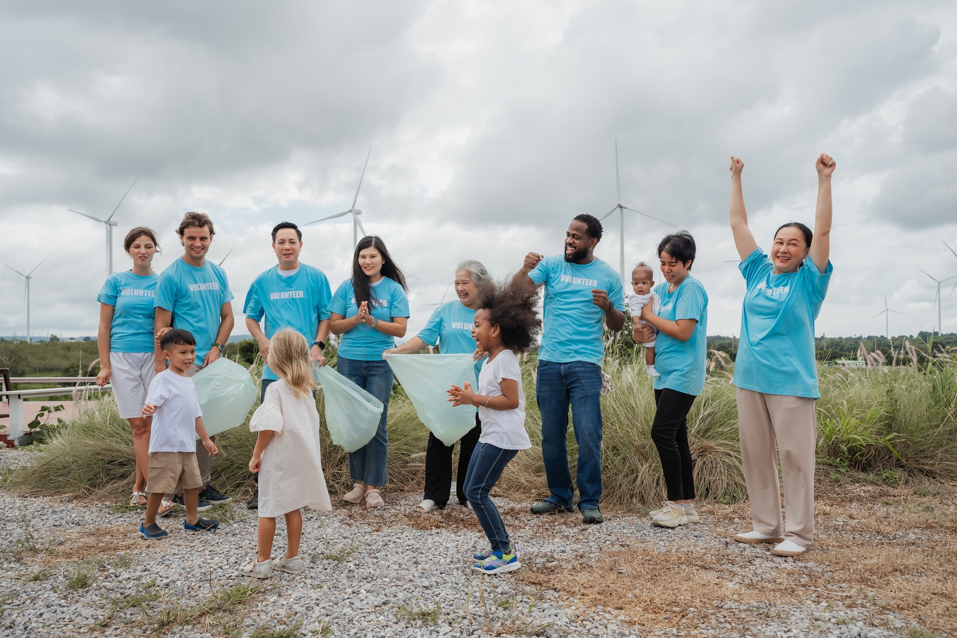 A group of people are standing on a hill, some of them are holding green bags. They are all wearing blue shirts. environment, clean, wind turbine, volunteer, community, teamwork, social, care, nature